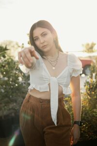 Stylish woman wearing white crop top and brown trousers posing outside in sunlight.