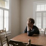 A man sits alone at a table in a bright room, displaying deep contemplation.